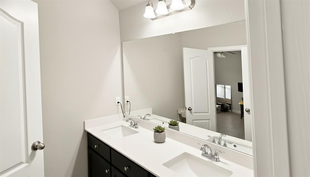 Double vanity featuring dark cabinetry, solid-surface countertop, dual integrated rectangular sinks, polished chrome faucets, and full-width vanity mirror with integrated three-bulb light fixture