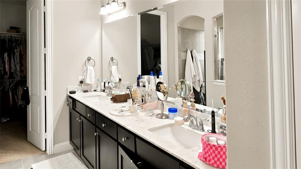 Double vanity featuring dark cabinetry, dual undermount sinks, light-toned stone-finish countertops, and a full-width vanity mirror with integrated lighting