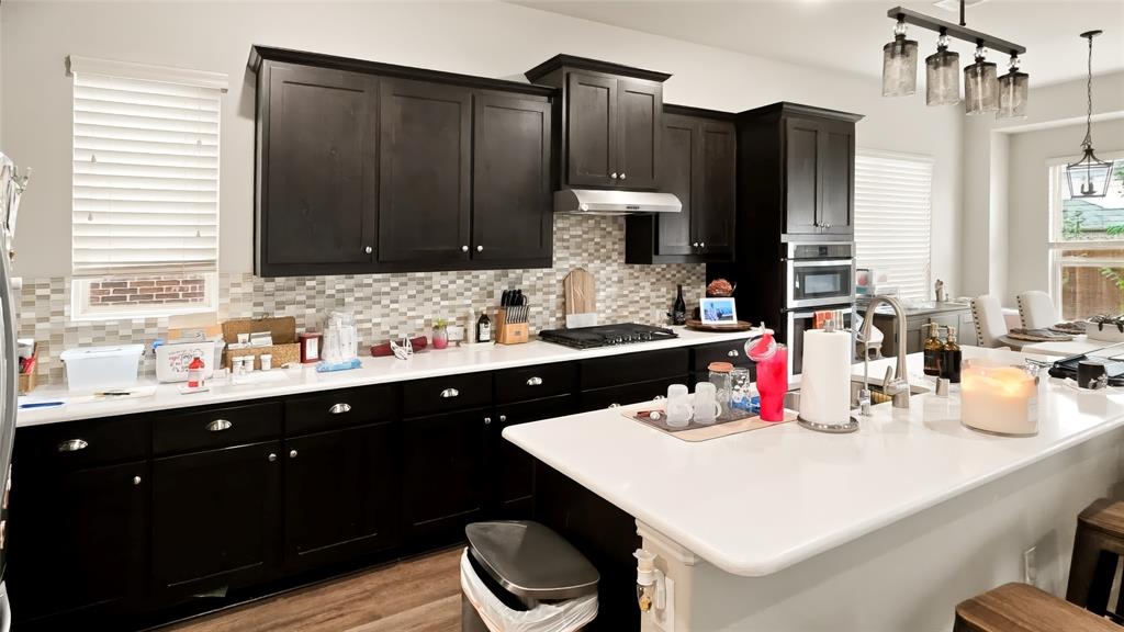 Kitchen featuring dark-finish cabinetry, white countertops, mosaic tile backsplash, built-in double wall ovens, and a center island with sink