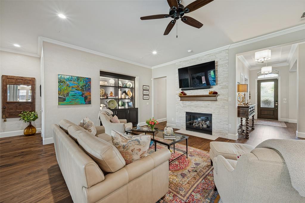 Living area featuring wood-finish flooring, a white stacked stone fireplace with a wood mantel, a ceiling fan, recessed lighting, and a built-in display cabinet with glass doors