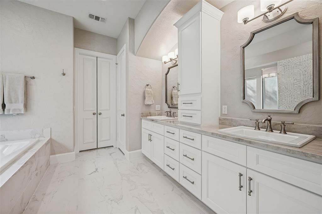 Dual vanity with white cabinetry and integrated sinks, stone-finish countertops, large soaking tub with stone-finish surround, built-in storage tower, and polished tile flooring