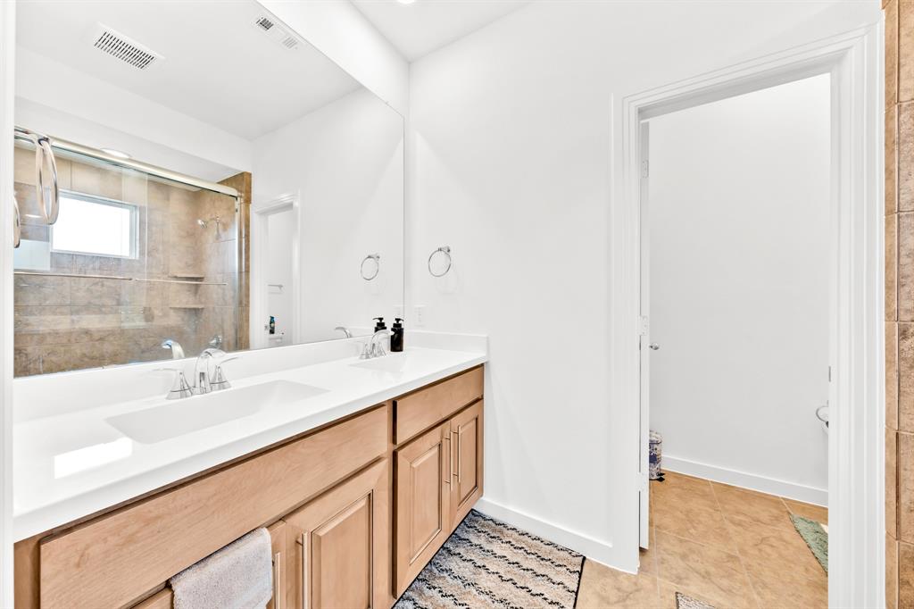 Double vanity with integrated sink, wood-finish cabinetry, frameless mirror, and glass-enclosed shower with tile surround