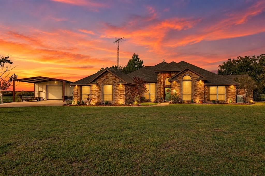 Stone masonry exterior featuring an arched entryway, shingle roof, expansive front lawn, and a metal-roofed detached structure