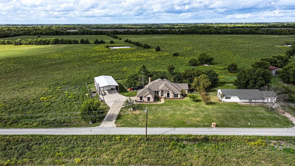 Aerial perspective of a multi-structure property featuring a main residence with a pitched roof, a detached metal-sided workshop, and a secondary structure with a covered porch, all situated on expansive acreage