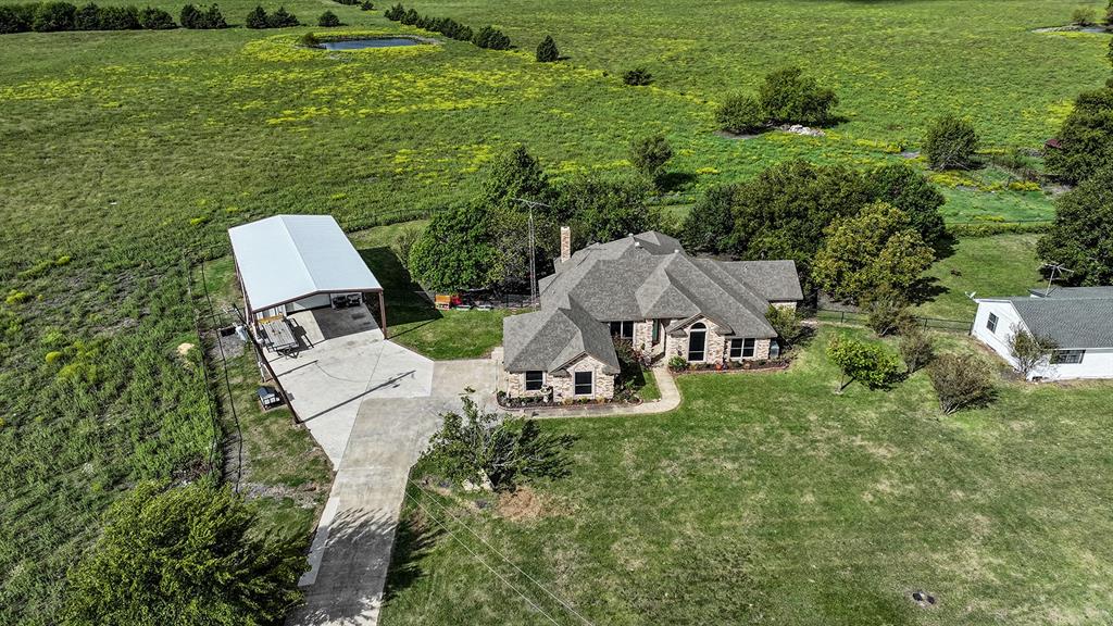 Aerial view of a sprawling property featuring a multi-gabled residence with stone masonry, an expansive concrete driveway, a detached metal-roofed carport, and fenced pasture grounds