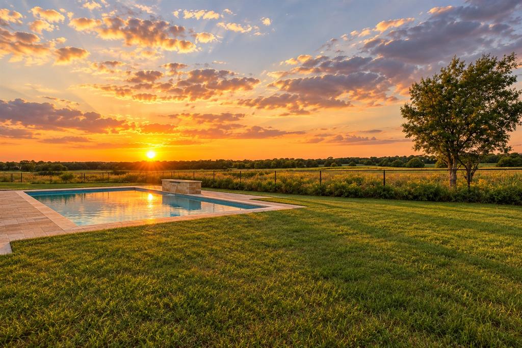 In-ground swimming pool featuring a stone-clad waterfall edge, paved deck, expansive lawn, and metal post fencing