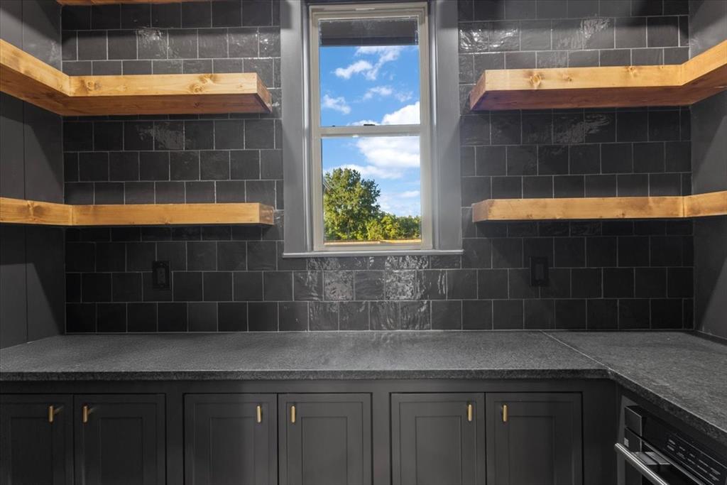 Built-in cabinetry with gold-finish hardware, dark stone countertops, dark tiled backsplash, and natural wood floating shelves