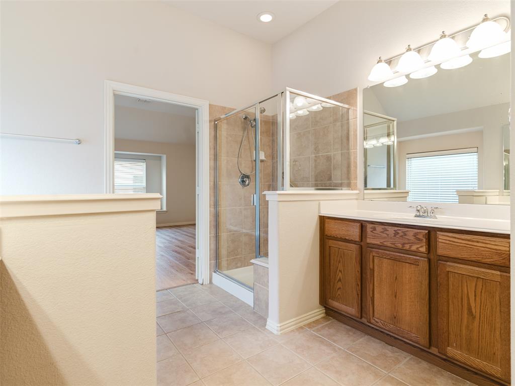 Bathroom featuring a wood-finish vanity with a solid-surface countertop, ceramic tile flooring, a glass-enclosed shower with tiled walls, and a large-format mirror with integrated lighting
