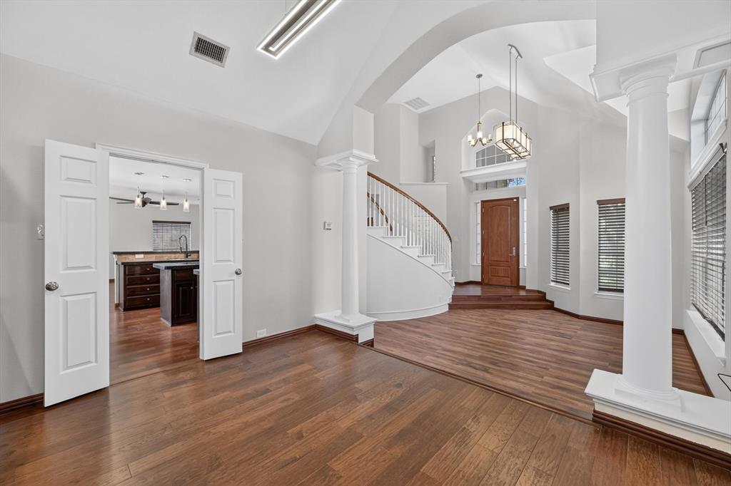 Open-concept floor plan featuring vaulted ceilings, wood-finish flooring, a curved staircase with a wooden handrail, classic structural columns, and a multi-paneled wood entry door