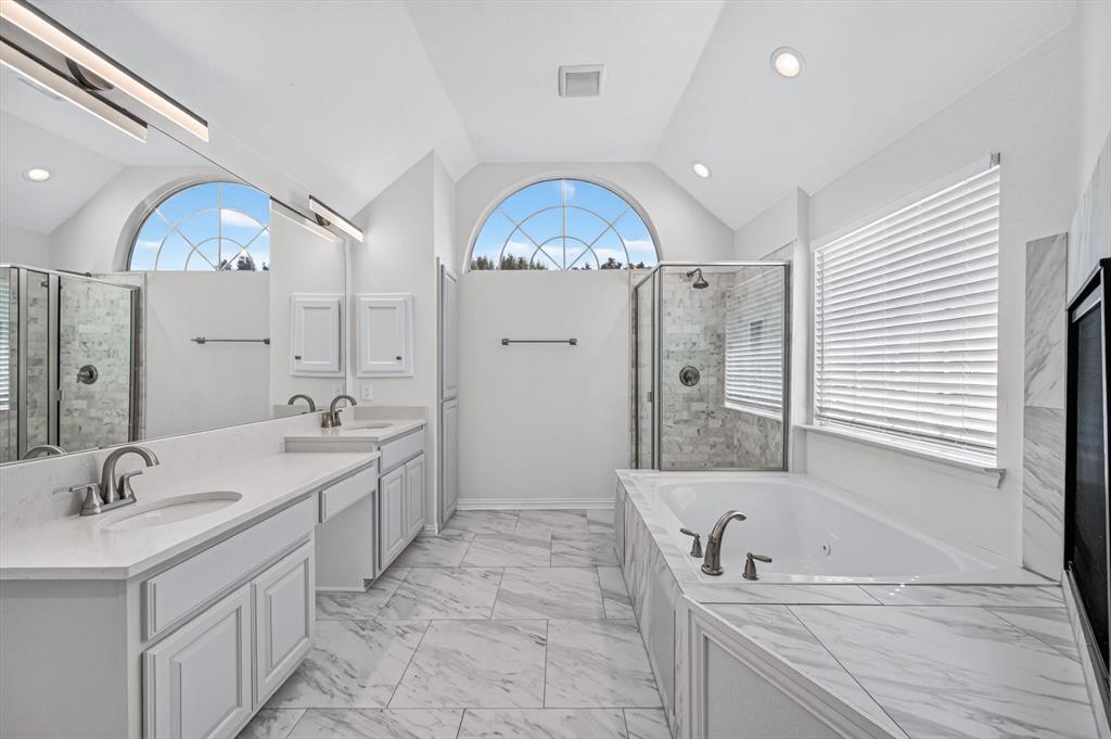 Bathroom featuring a dual-sink vanity with white cabinetry, quartz-surface countertops, arched window, soaking tub, and a glass-enclosed shower with stone-tile surround