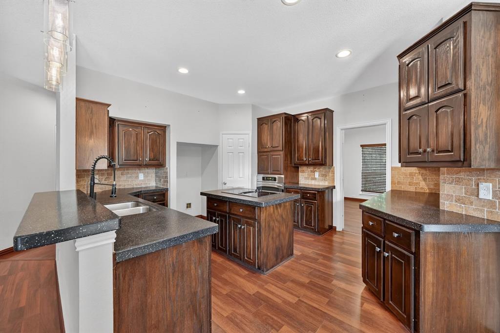 Kitchen featuring wood cabinetry, an island with an integrated cooktop, dark speckled countertops, brick-patterned tile backsplash, and wood-finish flooring