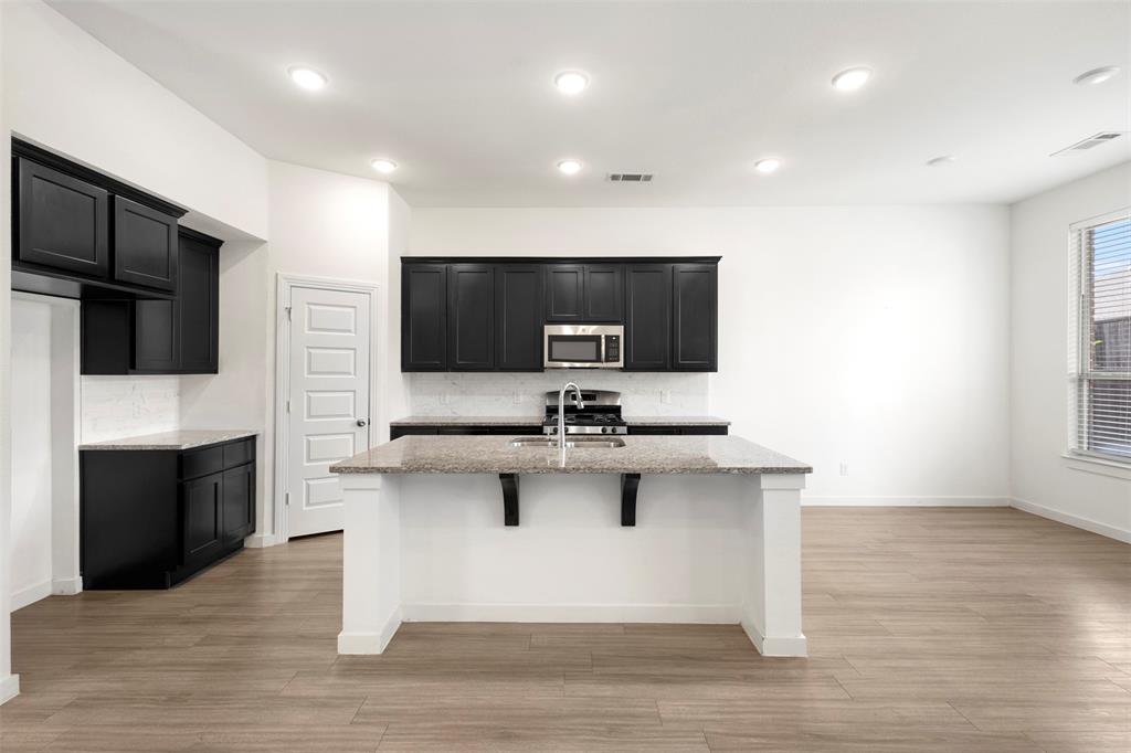 Kitchen featuring black shaker-style cabinetry, stone-surface center island with a stainless steel sink, wood-finish flooring, and recessed ceiling lighting