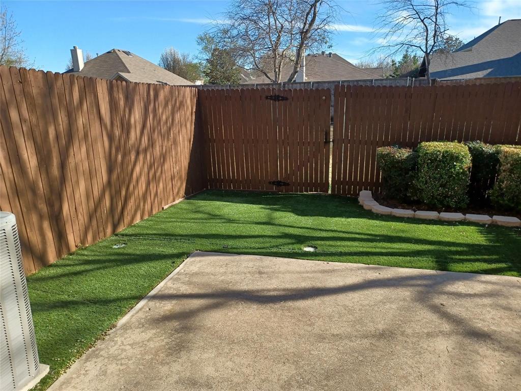 Fenced backyard featuring artificial turf, concrete patio, and landscaped shrubs with stone edging