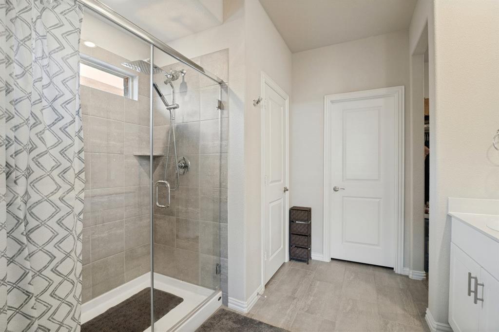 Walk-in shower with floor-to-ceiling tile surround, glass enclosure, and rainfall showerhead, complemented by wood-finish flooring and white vanity cabinetry with metallic pulls