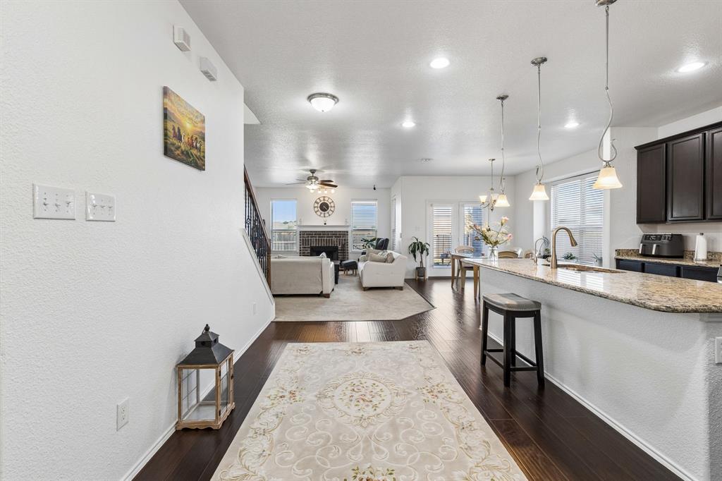 Dark wood-finish flooring extending into the living area, kitchen island with granite countertops and pendant lighting, dark wood cabinetry, and a living area featuring a fireplace with a stone surround