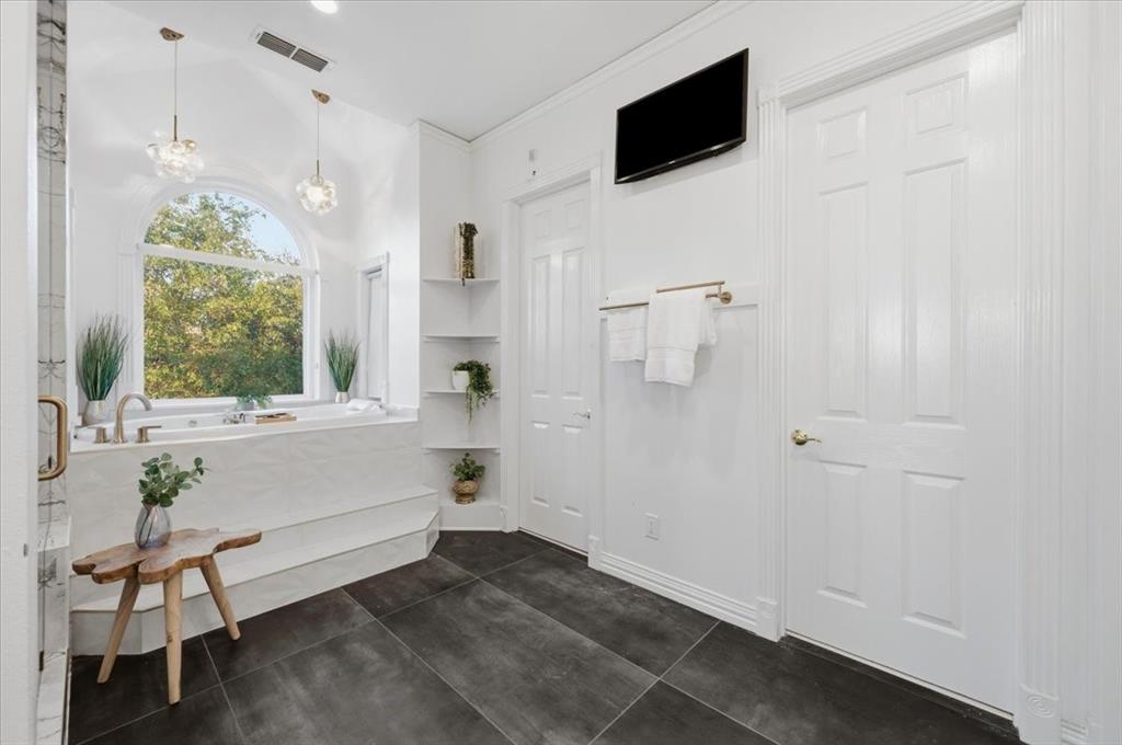Soaking tub with a tiled surround, arched window, dark tile flooring, built-in corner shelving, and dual glass pendant lighting