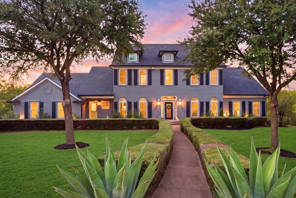 Two-story residence featuring a gray brick facade, dark window shutters, gabled rooflines, a central covered entryway, and mature landscape elements including manicured shrubbery and trees Two-story residence featuring a gray brick facade, dark window shutters, gabled rooflines, a central covered entryway, and mature landscape elements including manicured shrubbery and trees