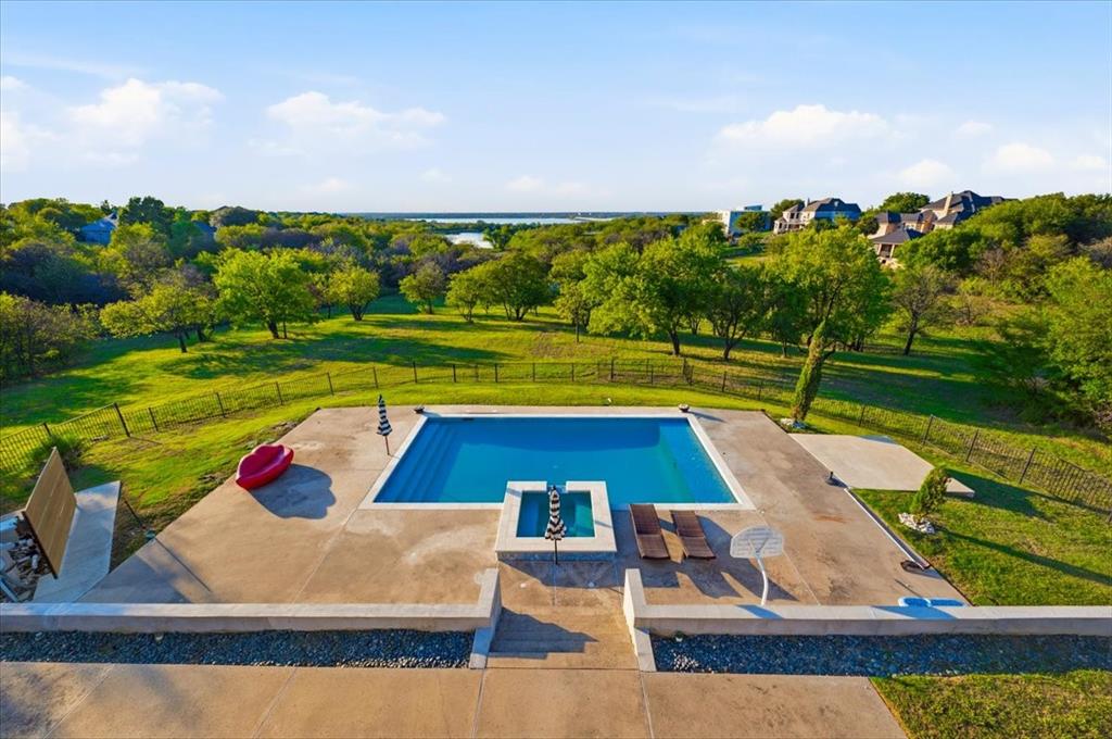 In-ground swimming pool with an attached spa, expansive concrete patio, metal perimeter fencing, and mature trees across a multi-acre landscape