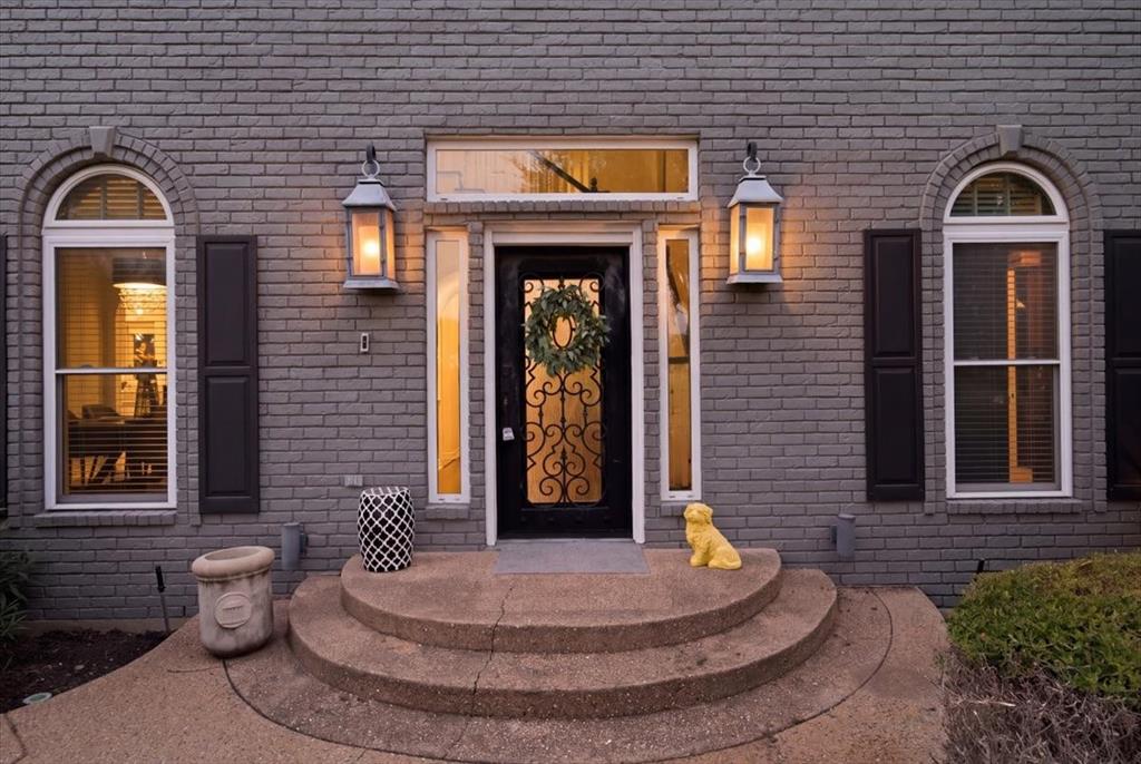 Gray brick exterior featuring a decorative iron-glass entry door, arched windows with shutters, dual exterior lanterns, and a curved concrete stoop with a transom window