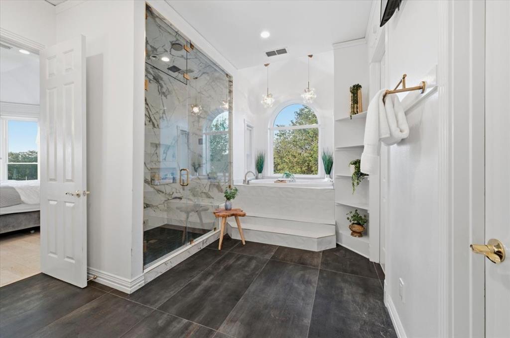 Walk-in shower with stone-finish tile surround and glass enclosure, deep soaking tub, arched window, and built-in shelving, complemented by dark large-format tile flooring