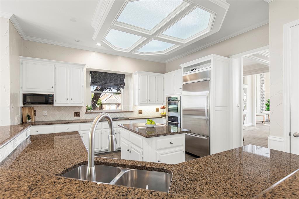 An expansive breakfast bar layout houses a double basin sink and a pull-down faucet, overlooking the bright cooking space with clean white cabinetry and an architectural ceiling light feature.