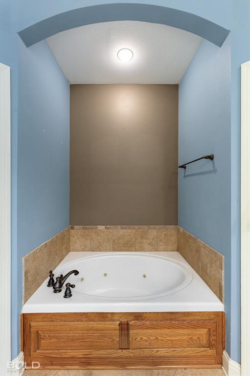 Jetted soaking tub featuring an arched recessed alcove, stone-finish tile surround, dark metal faucet hardware, and wood-finish cabinetry