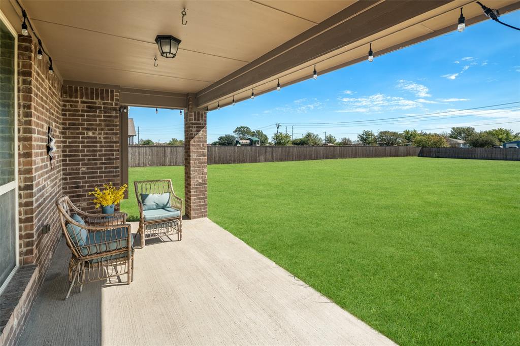 Covered concrete patio featuring brick exterior columns, exterior wall-mounted lighting, and an expansive level backyard enclosed by wood fencing