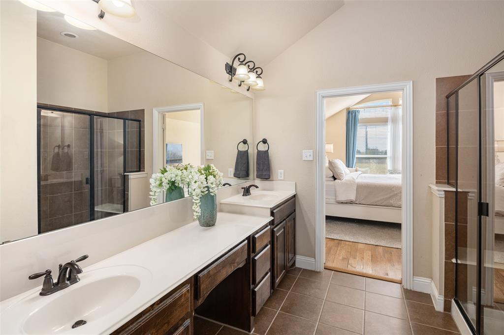 Dual-sink vanity with solid-surface countertop and dark-finish cabinetry, expansive wall-to-wall mirror, tiled flooring, and a glass-enclosed shower with tiled surround