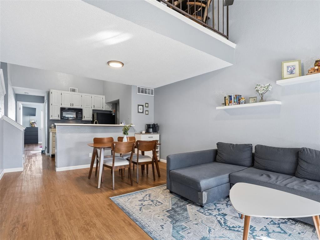 Open-concept area featuring wood-finish flooring, a lofted mezzanine with dark metal railing, white wall-mounted shelving, and a kitchen peninsula