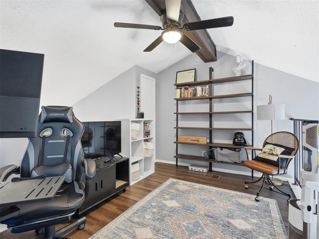 Sloped ceiling accented with a structural wood-finish beam, ceiling fan with integrated lighting, wall-mounted industrial-style shelving, and wood-finish flooring