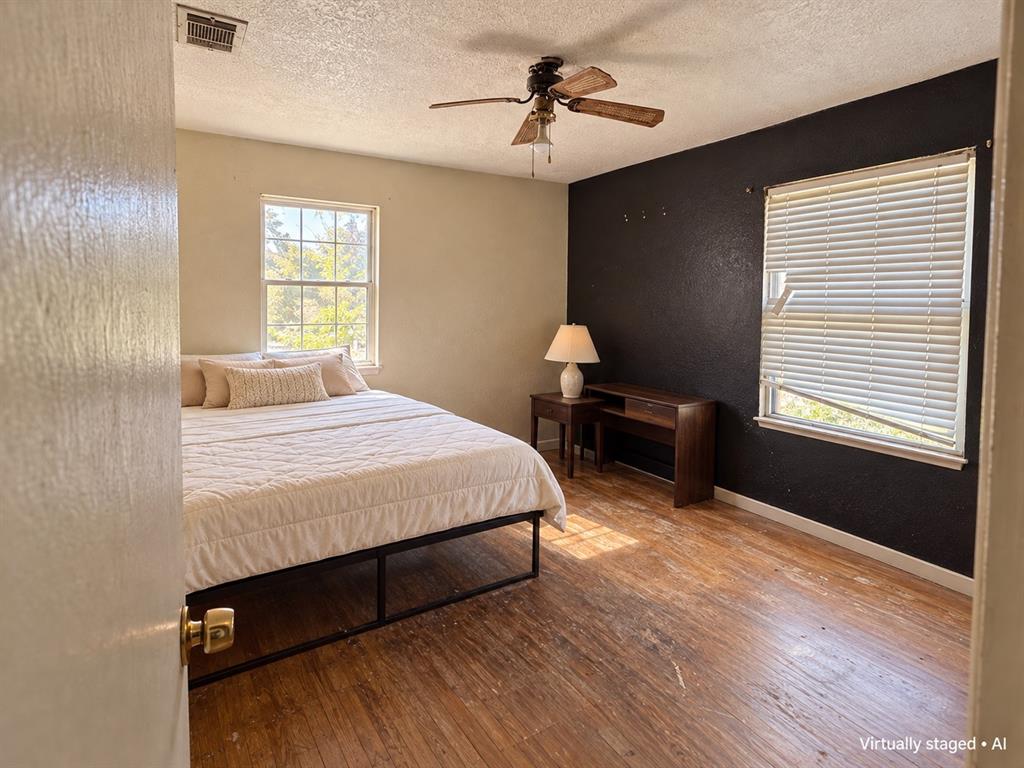 Bedroom featuring wood flooring, contrasting accent wall, ceiling fan, and two windows providing natural light. Virtually staged.