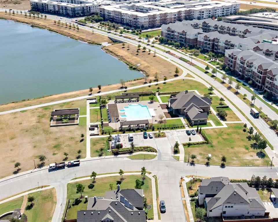 Aerial perspective of the community recreation complex featuring a swimming pool, covered pergola structures, a clubhouse building, and paved parking area, situated along a large pond