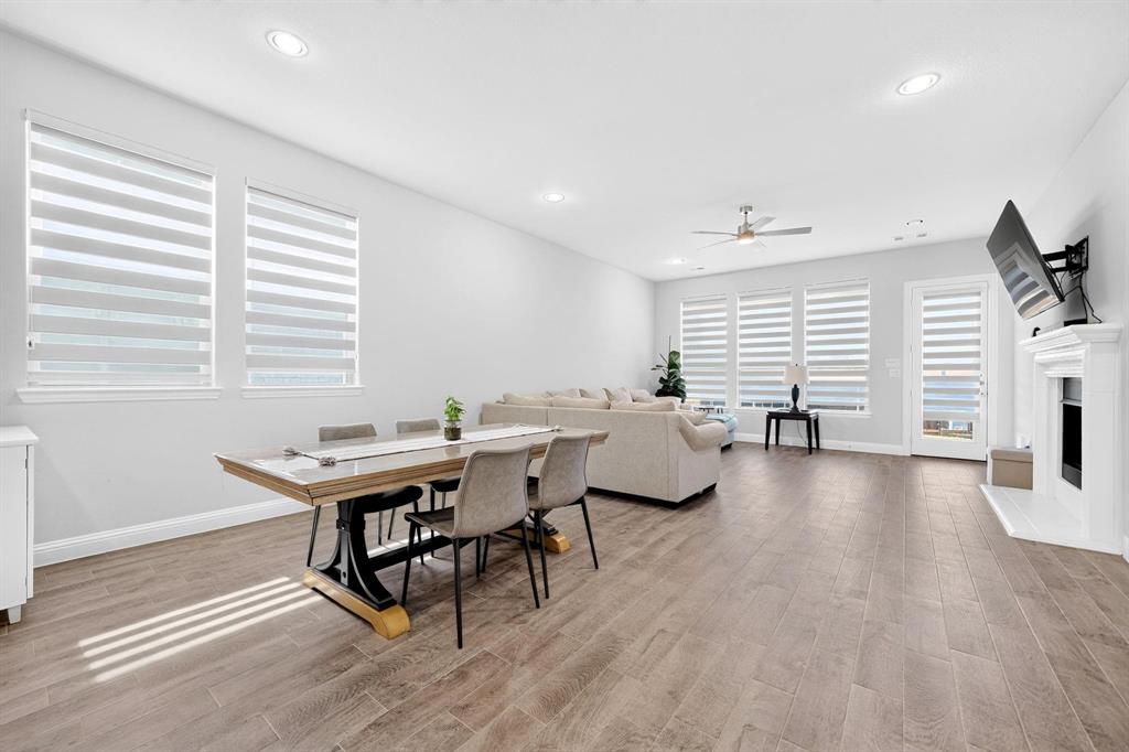Open-concept space featuring wood-finish flooring, recessed lighting, a ceiling fan, tiered window shades, and a fireplace with a white mantel