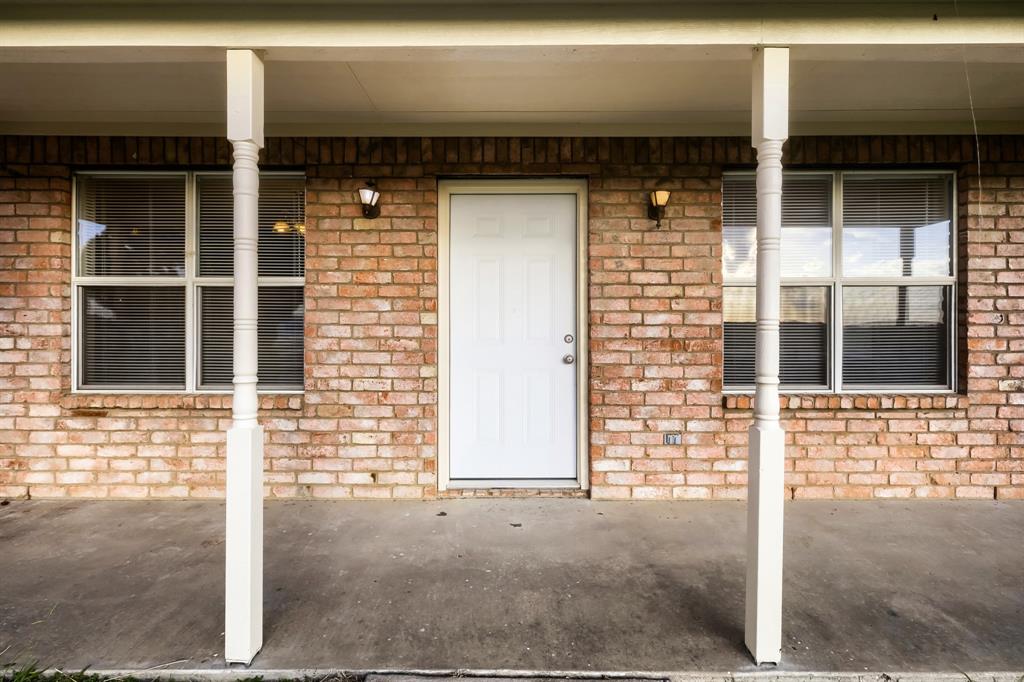 Covered front porch with concrete flooring, brick exterior wall, dual white support columns, and two wall-mounted light fixtures