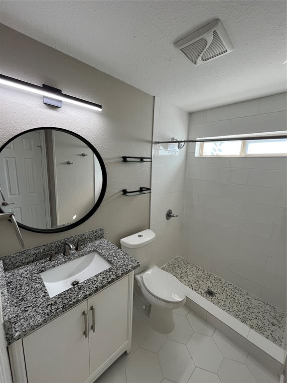 Bathroom featuring a stone-composite countertop, white vanity cabinet with metallic hardware, circular wall-mounted mirror, and a walk-in shower with white tile surround and pebble-texture flooring