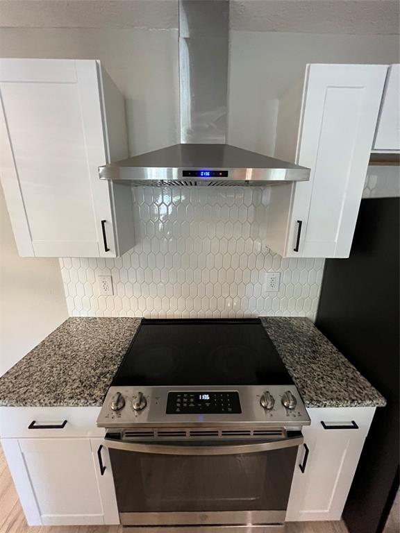 Stainless steel range, chimney-style range hood, white shaker-style cabinetry, stone-finish countertops, and white elongated hexagonal tile backsplash