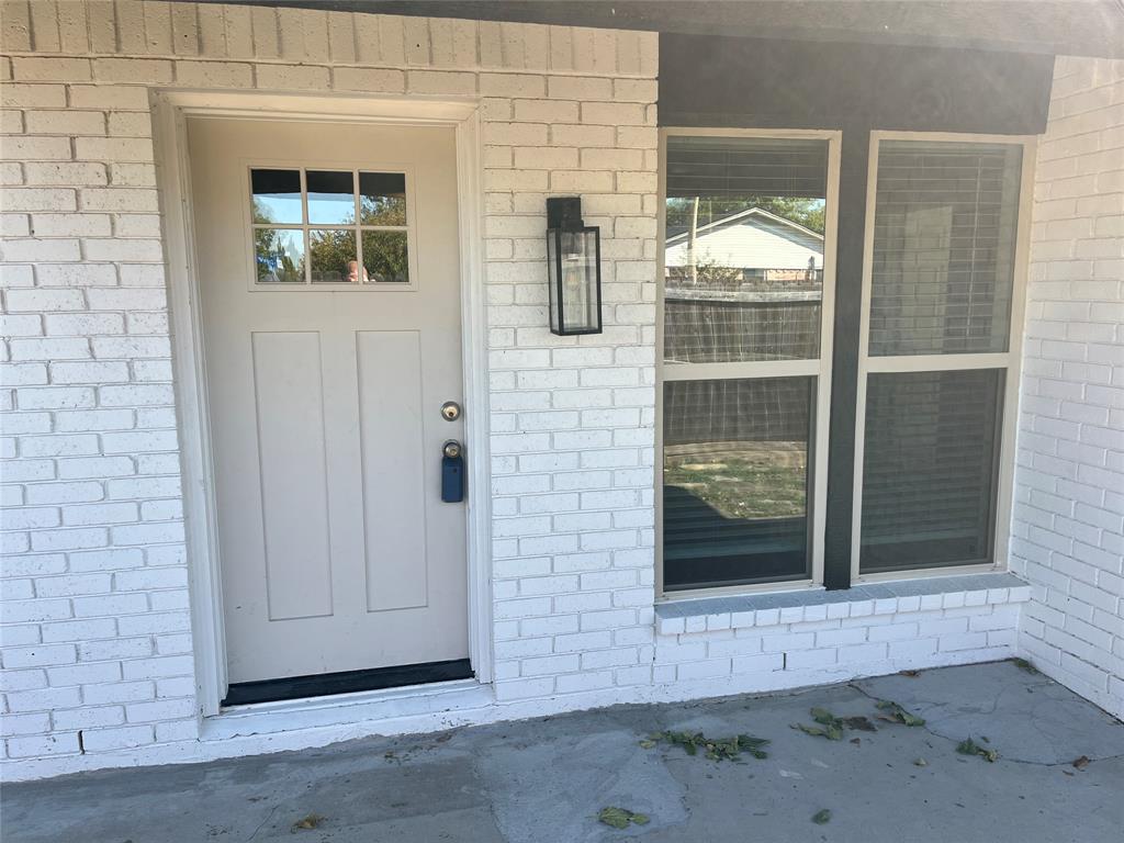 White brick exterior featuring a single-panel front door with a six-pane window insert, paired with dual windows and a rectangular matte-black wall-mounted lantern