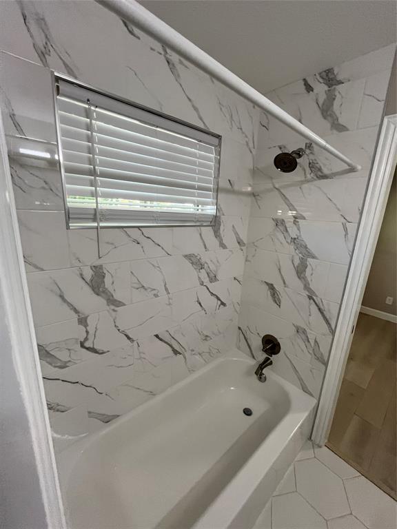 Bathtub enclosure featuring large-format patterned tile walls, a built-in shower head, a bronze-finish faucet, and an integrated window with horizontal blinds