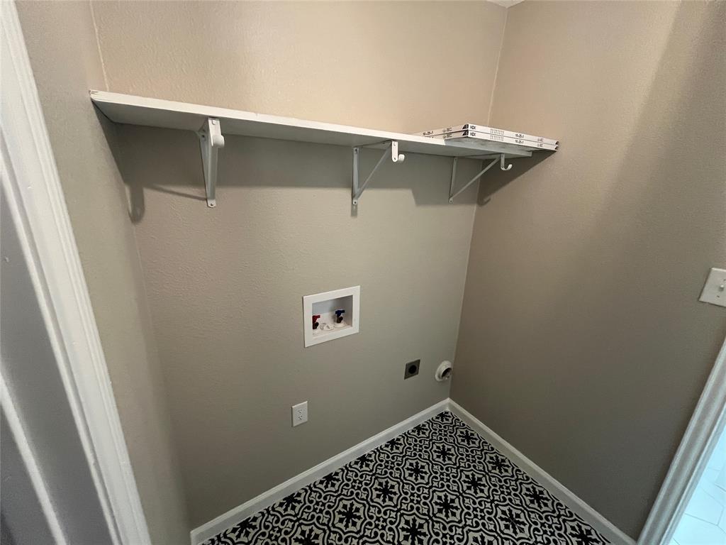 Utility area featuring patterned ceramic tile flooring, a wall-mounted shelving unit with support brackets, and recessed plumbing hookups