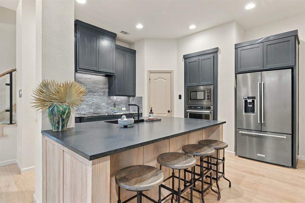 Kitchen featuring dark-toned cabinetry, a spacious island with a solid-surface countertop, stone-finish backsplash, built-in stainless steel appliances, and wood-finish flooring