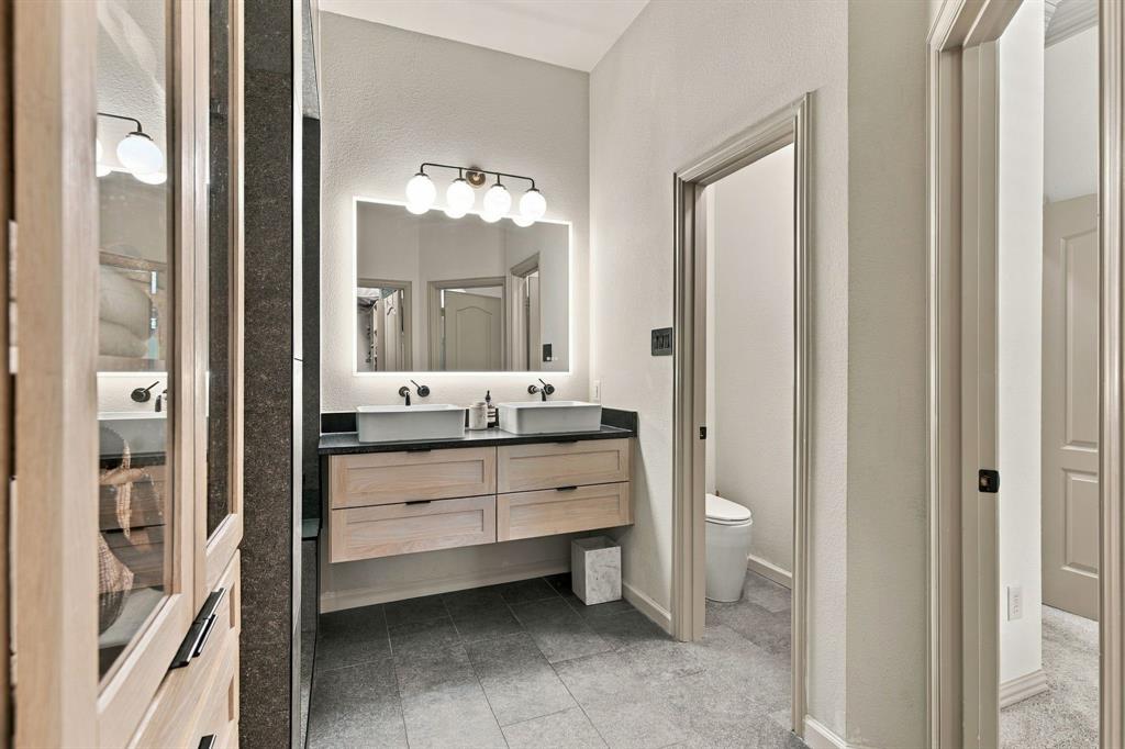 Floating dual vanity with light-tone wood cabinetry, rectangular vessel sinks, dark countertop, backlit vanity mirror, and slate-finish tile flooring