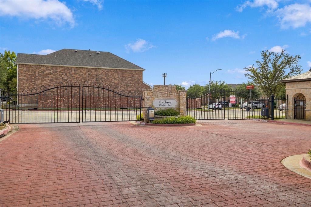 Gated entrance featuring red brick paver driveway, ornate wrought-iron security gate, stone monument signage, and perimeter brick wall