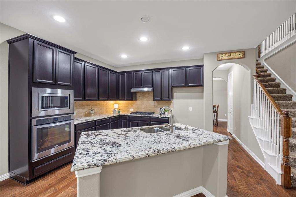Kitchen featuring dark cabinetry, stone countertops, stainless steel double wall ovens, a central island with a double-basin sink, and recessed lighting