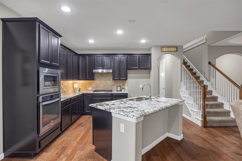 Kitchen featuring 42-inch dark cabinetry, granite countertops, stainless steel built-in oven and microwave, center island with a dual-basin sink, and wood-finish flooring