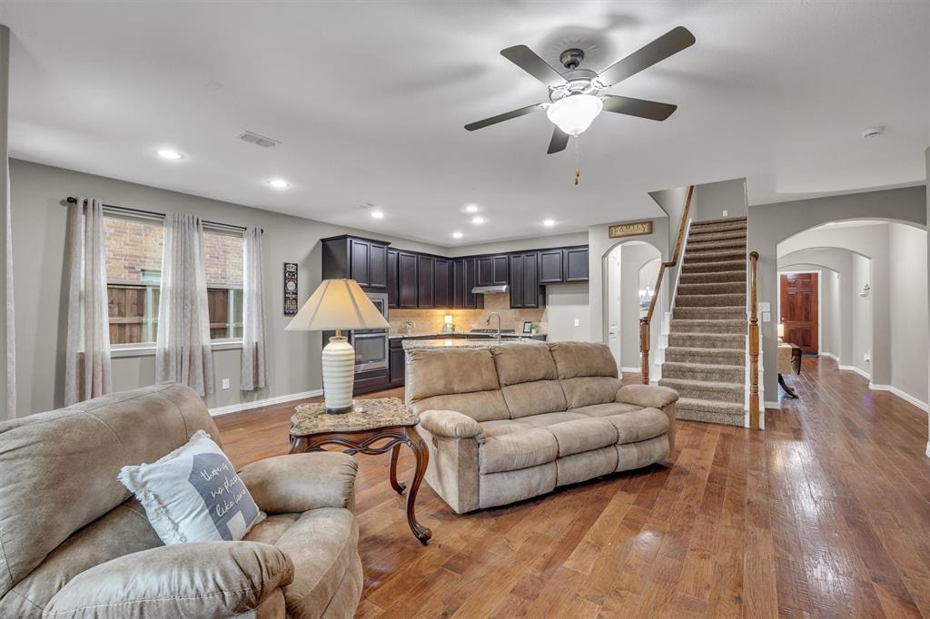 Open-concept interior featuring wood-finish flooring, recessed lighting, dark cabinetry with stone countertops, a ceiling fan with integrated lighting, and a staircase with a wooden handrail