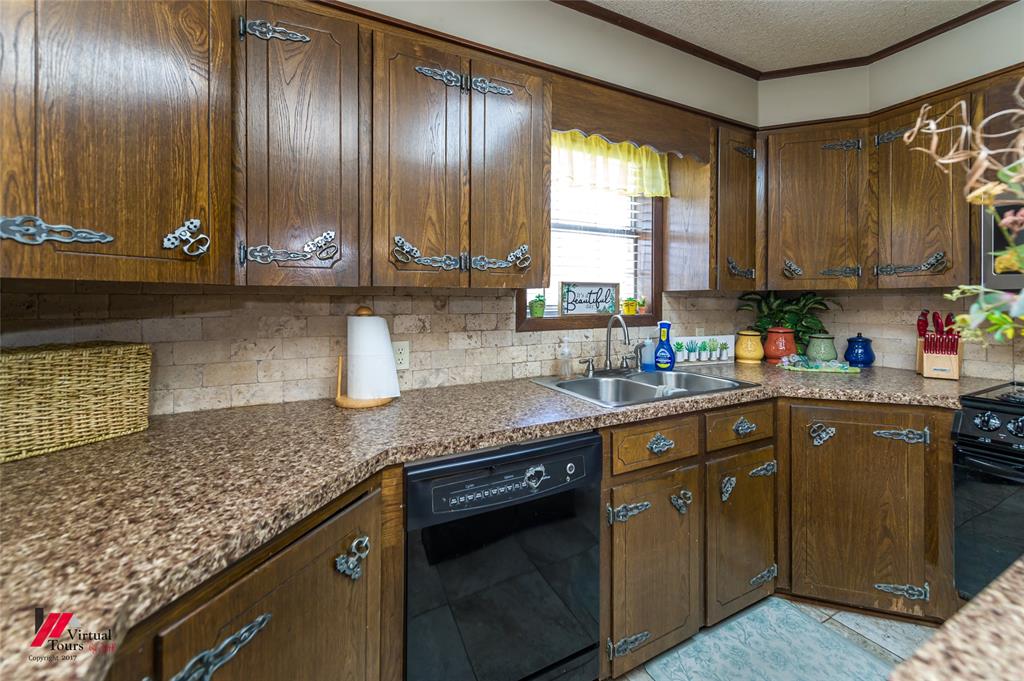 Custom stained wood cabinetry featuring decorative metal hardware, mottled stone-finish countertops, natural stone tile backsplash, integrated stainless steel double-basin sink, and dark ceramic tile flooring