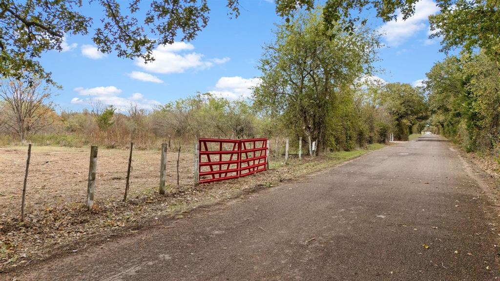 Expansive acreage featuring wire fencing, a metal gate, mature trees, and frontage on a paved road