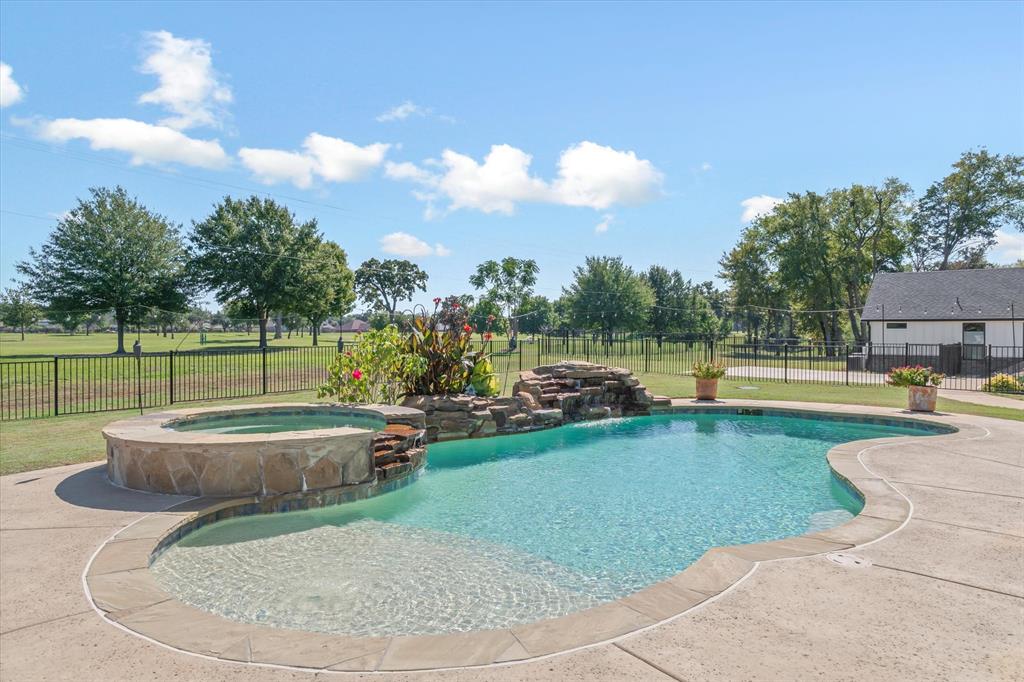 In-ground pool featuring a stone waterfall, integrated raised spa, concrete decking, and metal perimeter fencing