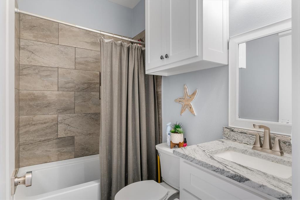 Bathroom featuring a vanity with a light-toned stone countertop, white cabinetry, a framed mirror, and a tub surround finished in large-format neutral-toned tiles