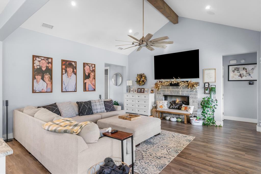 Vaulted ceilings with exposed wooden beam, stacked-stone fireplace, wood-finish flooring, and multi-blade ceiling fan